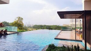 A rooftop infinity pool with a wooden deck and lounge chairs, surrounded by greenery and city buildings. - Delight Homestay Mosaic Southkey (Plentong)