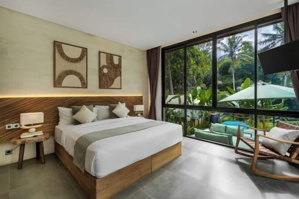A modern bedroom with a large bed, a wooden headboard, a nightstand, and a view of lush greenery through large windows.