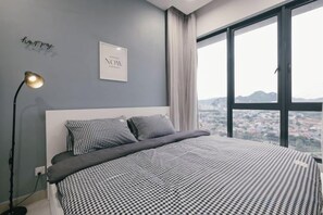A bedroom with a large bed, a bedside lamp, a view of a cityscape through a window, and a wall with a framed text. - Mini House Gombak2BR 5paxAyuman Suites (Setapak)