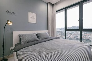 Ein Schlafzimmer mit einem großen Bett, einem Nachttisch, einer Lampe und Blick auf eine Stadtlandschaft durch große Fenster.