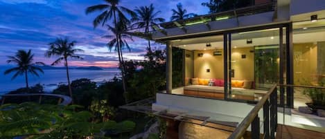 A modern house with a glass balcony overlooking a tropical landscape at dusk.