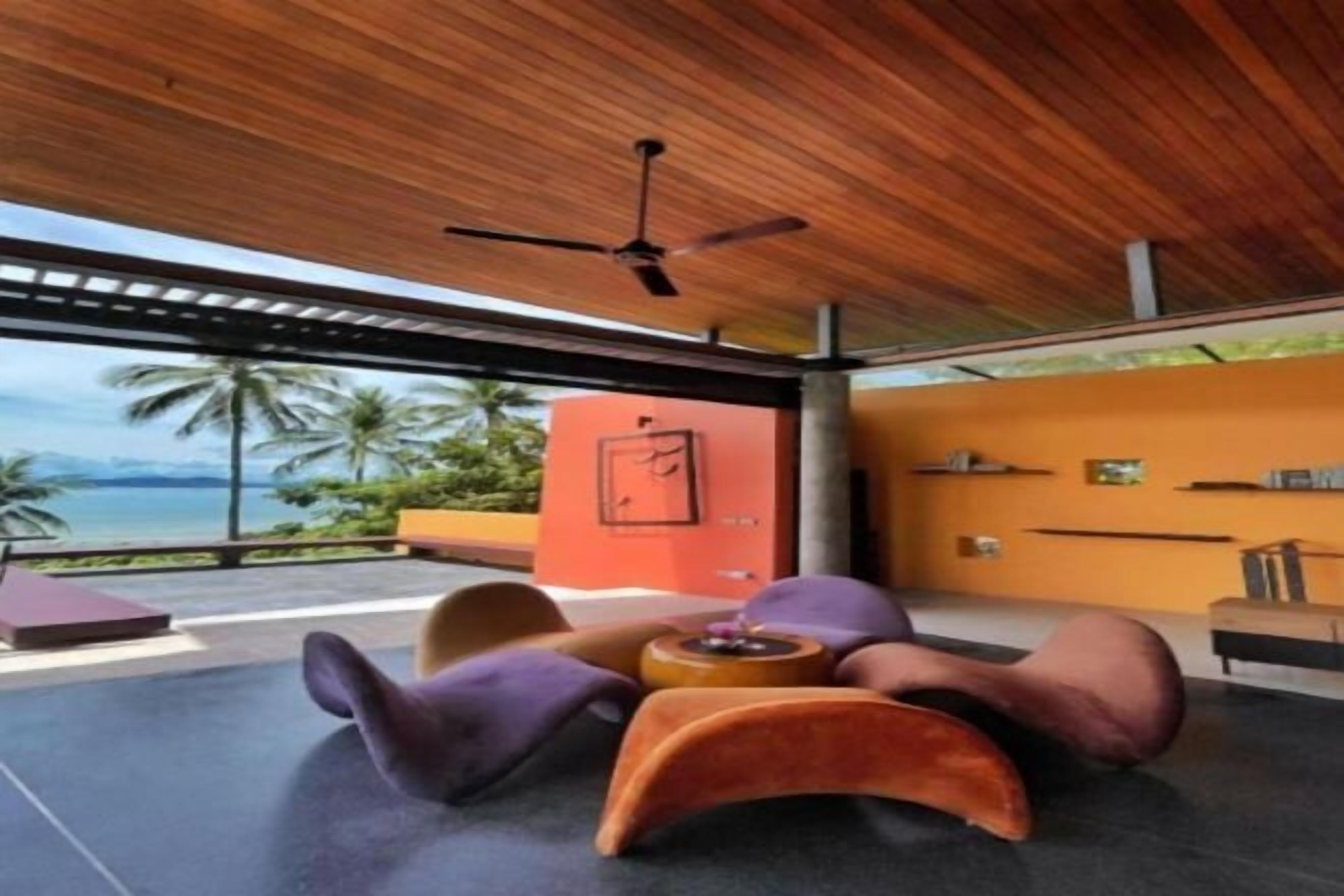 A modern room with a wooden ceiling and a view of the ocean. There are colorful chairs and a small table with a vase.
