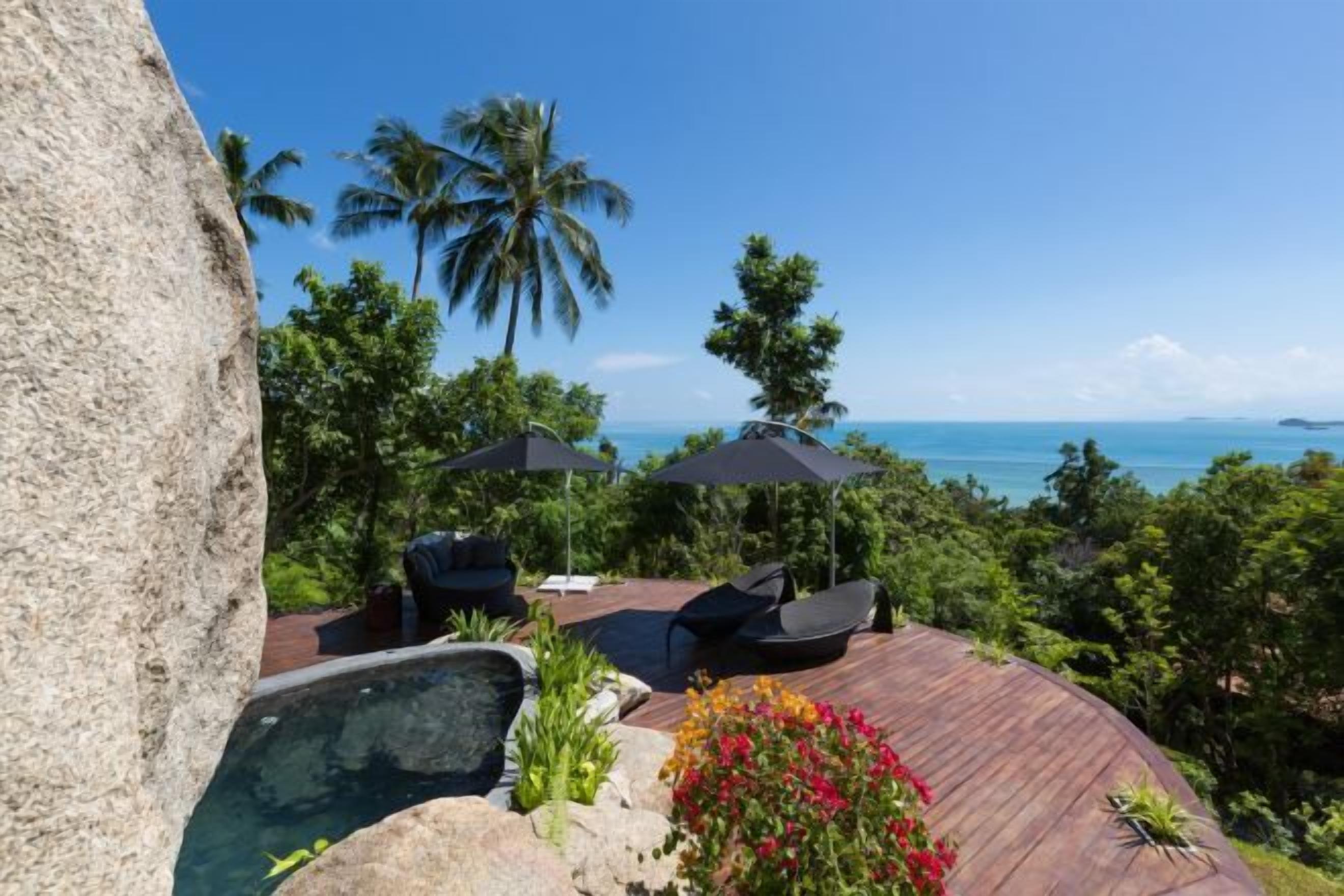 Una terraza de madera con jacuzzi, sillas de descanso y sombrillas, con vista a una playa con palmeras y el océano.