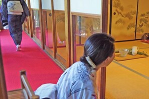 Un interno tradizionale giapponese con pavimento in tatami, un tavolino con una tazza bianca e una piccola ciotola, e una persona che cammina lungo un corridoio.
