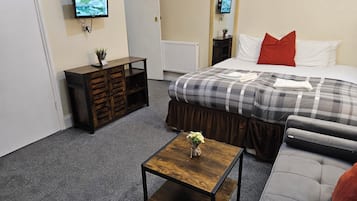Une chambre d’hôtel avec un lit, un canapé, une petite table en bois sur laquelle est posé un vase, une commode et un téléviseur fixé au mur.