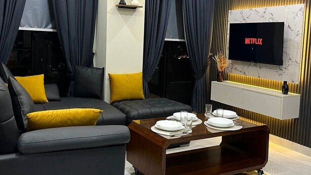 A modern living room with a sectional sofa, a coffee table, and a flat-screen TV mounted on a wall with a marble-like design. - Cozy Apartment in Bahria Town. (Rawalpindi)