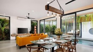 A modern living room with a glass dining table, orange sofa, and a TV. - VILLA ELIZA CANGGU (Bali)