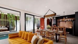 A modern living room with a yellow sofa, dining table, and chairs, a kitchen area, and a large window with hanging plants. - VILLA ELIZA CANGGU (Bali)
