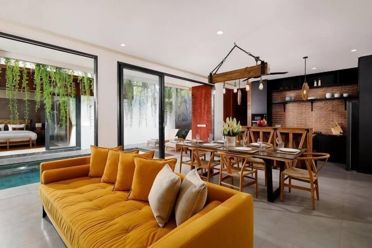 Un salon moderne avec un canapé jaune, une table à manger et des chaises, un coin cuisine et une grande fenêtre ornée de plantes suspendues.