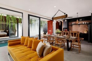 Un salon moderne avec un canapé jaune, une table à manger et des chaises, un coin cuisine avec un mur de briques apparentes et de grandes fenêtres ornées de plantes d’intérieur.