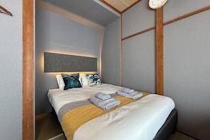 A neatly made bed with towels, a headboard with a light panel, and wooden paneling on the walls. - Nestay villa Tokyo Ueno (Tokyo)
