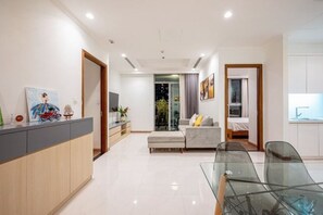 A modern living room with a glass dining table, a sofa, and a kitchen area visible through an open door. - An Corner Apt - Vinhomes Central Park High Floor Nice View and Free Pick up for 5 Nights (Ho Chi Minh City)