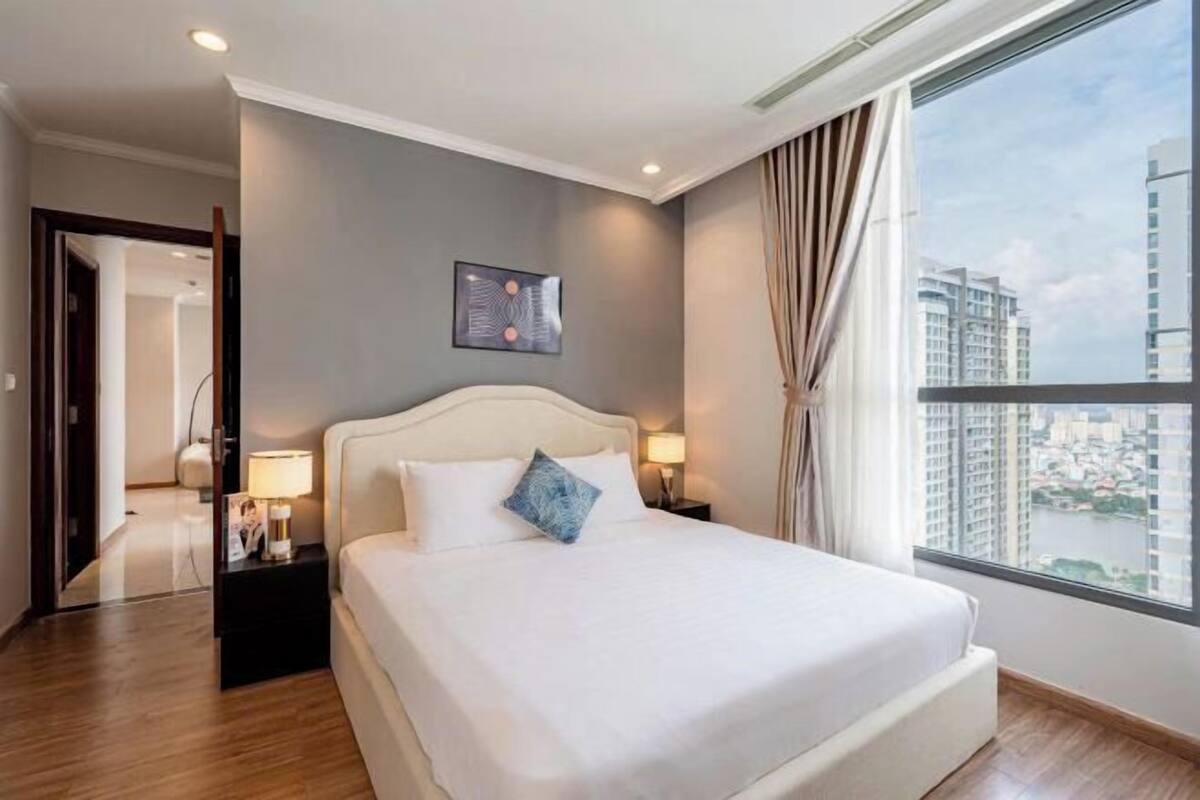 Ein modernes Hotelzimmer mit einem großen Bett, einem Nachttisch, einer Lampe, einem gerahmten Bild an der Wand und Blick auf die Stadt durch das Fenster.