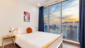 A modern hotel room with a large bed, a nightstand, a lamp, a painting, and a city view. - An Corner Apt - Vinhomes Central Park High Floor Nice View and Free Pick up for 5 Nights (Ho Chi Minh City)