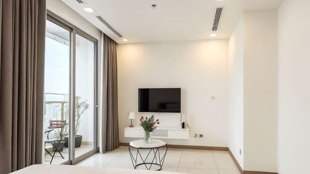 A modern hotel room with a large bed, a TV mounted on the wall, a small round table with a vase of flowers, and a balcony with a chair and potted plant. - An Corner Apt - Vinhomes Central Park High Floor Nice View and Free Pick up for 5 Nights (Ho Chi Minh City)