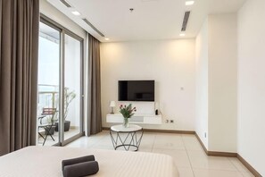 A modern hotel room with a large bed, a TV mounted on the wall, a small round table with a vase of flowers, and a balcony with a chair and potted plant. - An Corner Apt - Vinhomes Central Park High Floor Nice View and Free Pick up for 5 Nights (Ho Chi Minh City)