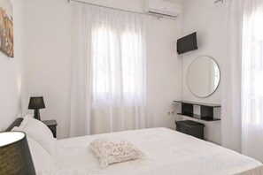 A bedroom with a bed, a bedside table, a lamp, a mirror, and a window with curtains. - Akakia Residence (Naxos)