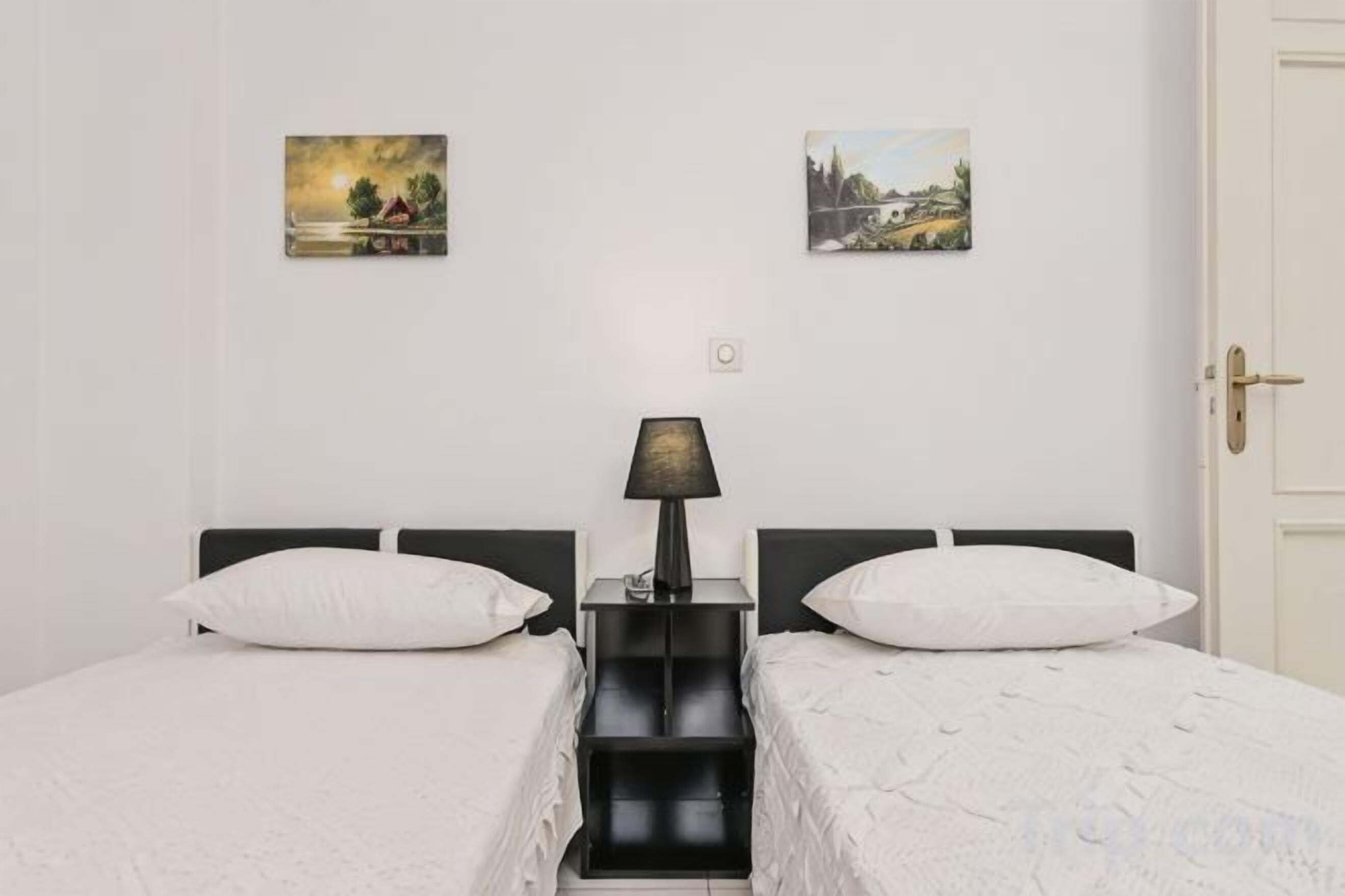A double bed with white bedding, a black nightstand with a lamp, and two wall-mounted paintings.