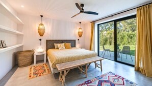 Ein Schlafzimmer mit einem Bett, Nachttischlampen, einem Deckenventilator, einer Bank, einem Teppich und einem großen Fenster mit Vorhängen.