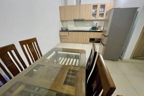 Un comedor con mesa de vidrio y sillas de madera, una cocina con armarios de madera clara y un refrigerador, además de un piso con baldosas.