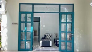 A room with a bed, a small table, and a chair. There is a door with a sign above it. - Nhà Nguyên Căn Costis 5 Phòng Ngủ (Phu Quoc Island)