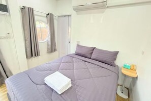 A bedroom with a bed, a bedside table, a window with curtains, and an air conditioning unit.