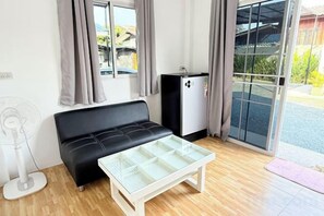A room with a black sofa, a small white table, a white fan, and a refrigerator. - Cina Home (Chiang Mai)