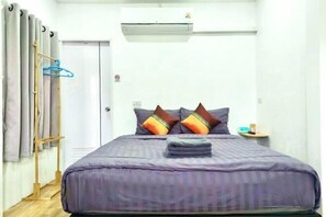 A bed with a purple bedspread, a wooden ladder, a small table with a lamp, and a door with a hanging garment rack. - Cina Home (Chiang Mai)