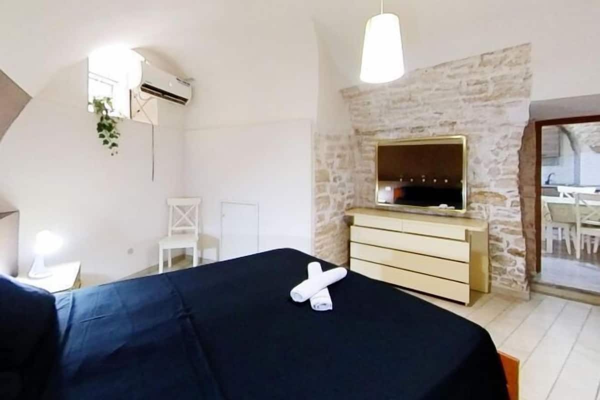 Une chambre à coucher avec un lit recouvert d’un drap bleu foncé, une commode en bois, un téléviseur fixé à un mur en pierre et une chaise blanche.