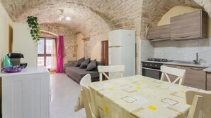 Een knus woongedeelte met een stenen plafond, een kitchenette, een eethoek met tafel en stoelen, en een bank.
