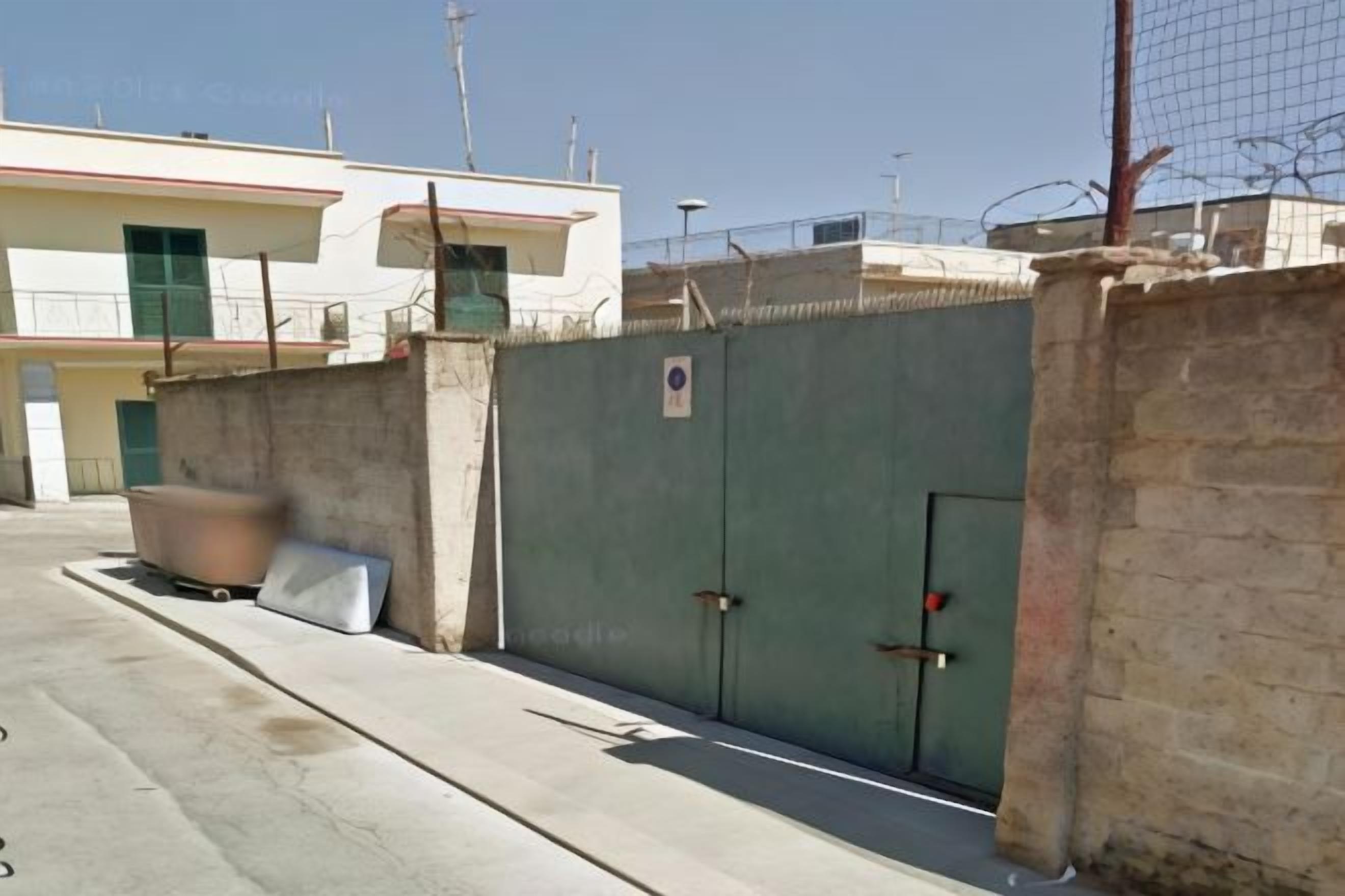 A gated area with a metal gate, a concrete wall, and a building with a flat roof in the background.