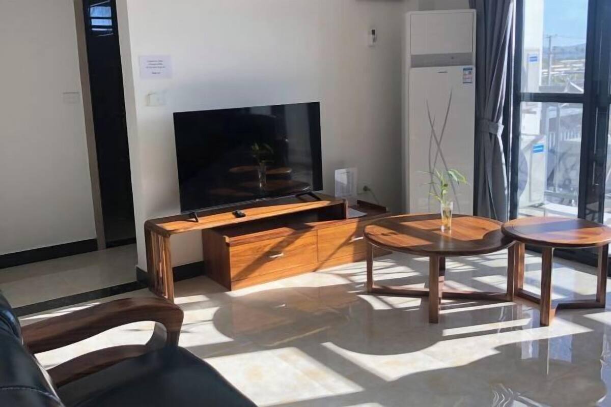 A modern living room with a flat-screen TV, wooden furniture, and a view through a large window.
