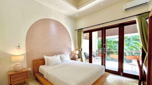 A hotel room with a large bed, a nightstand, a lamp, and a view of the outdoors through a sliding glass door. - Cove The Ricks (Bali)