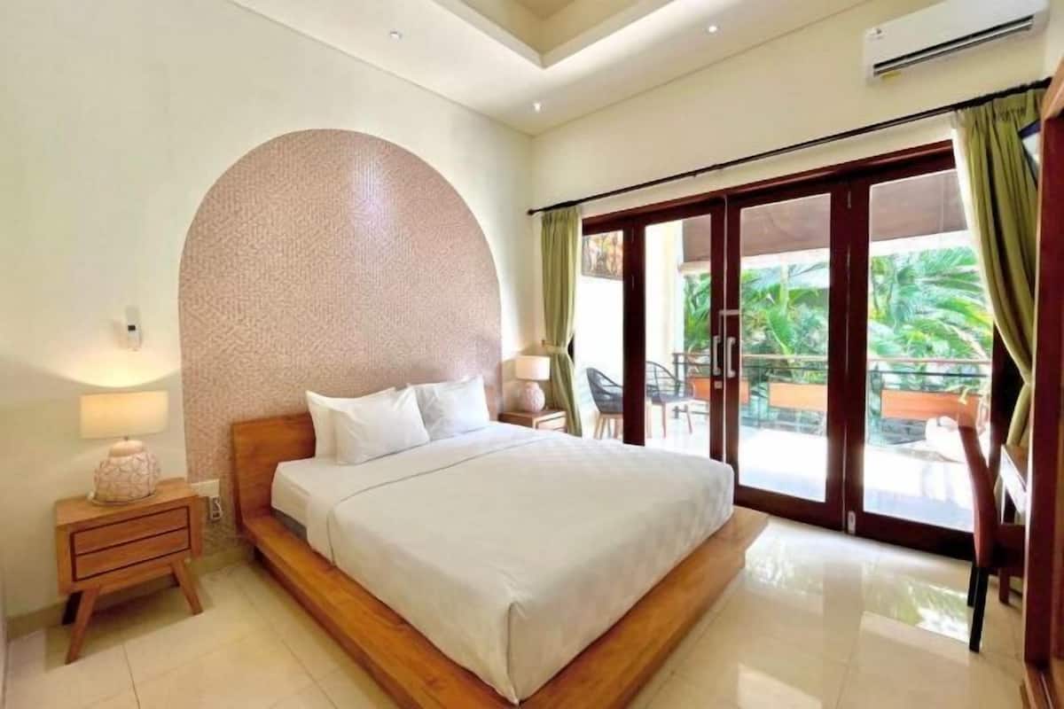 A hotel room with a large bed, a nightstand, a lamp, and a view of the outdoors through a sliding glass door.
