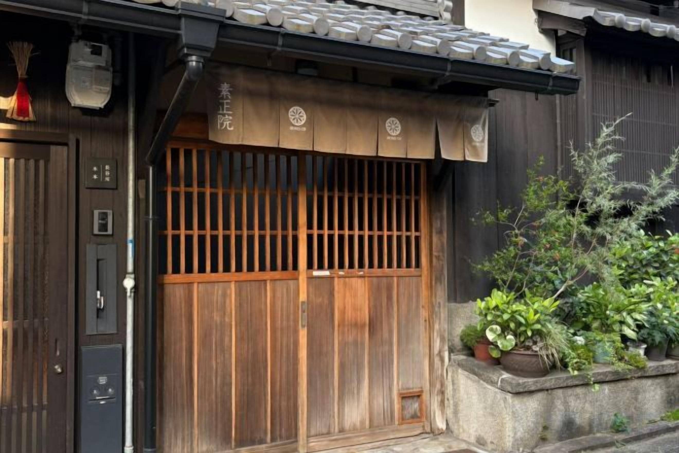 Edifício tradicional japonês com porta de correr de madeira, telhado telhado e pequena área de jardim com plantas.
