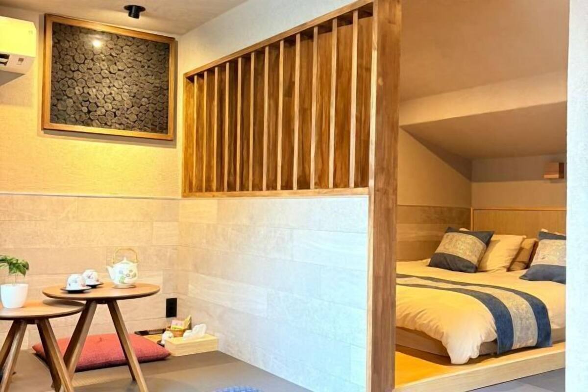 A bedroom with a bed, a small table with a teapot, a potted plant, and a wooden slat partition.