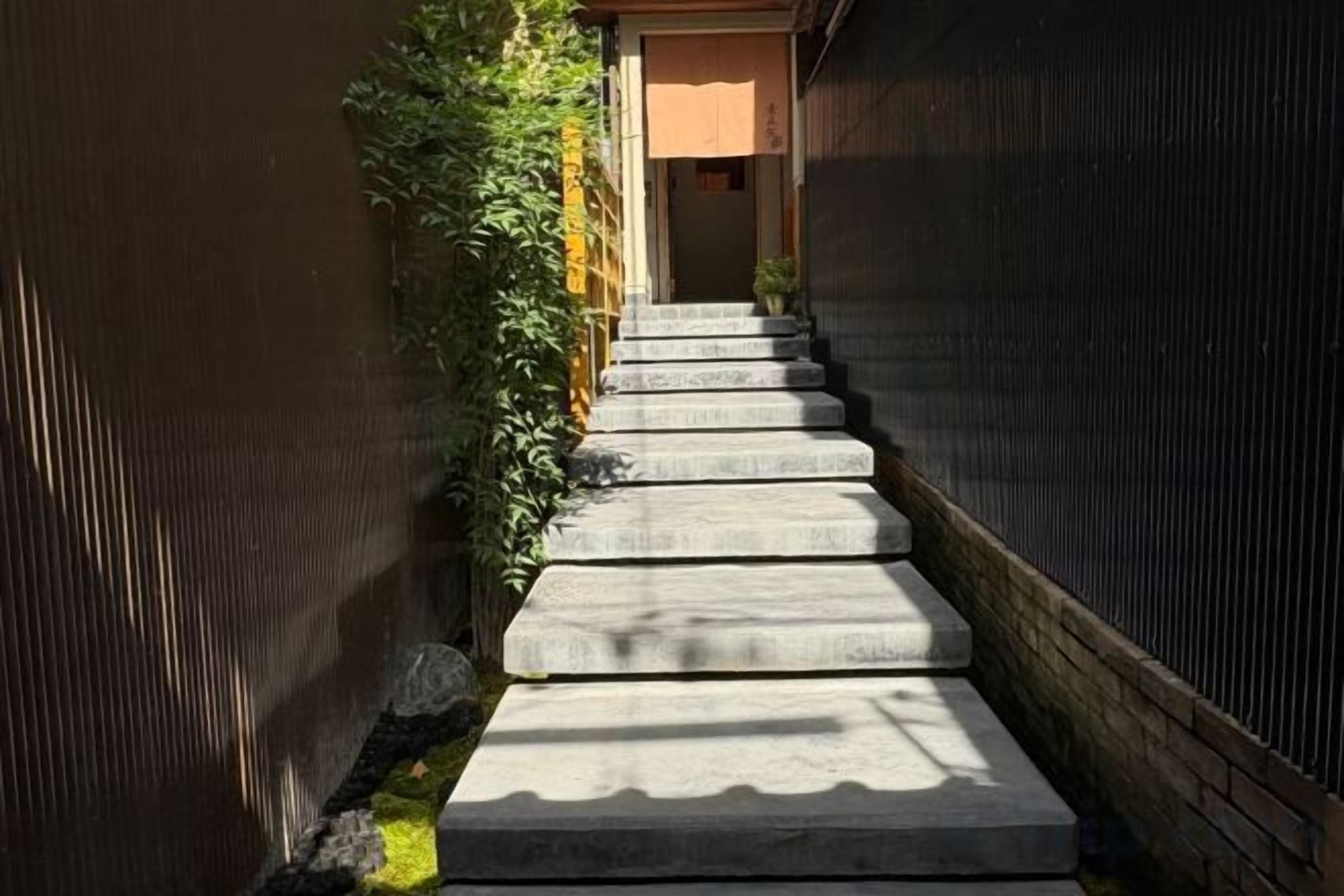 Escadaria de concreto que leva a uma porta, cercada por uma cerca de madeira e vegetação.