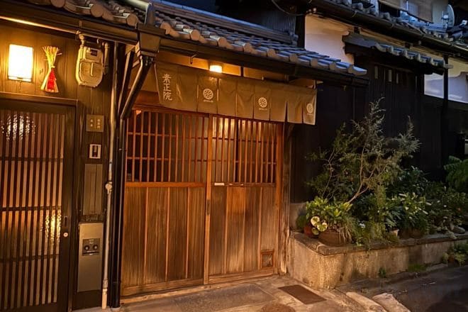 Um edifício tradicional japonês com portão de madeira, telhado de telhas e uma pequena área de jardim com plantas.