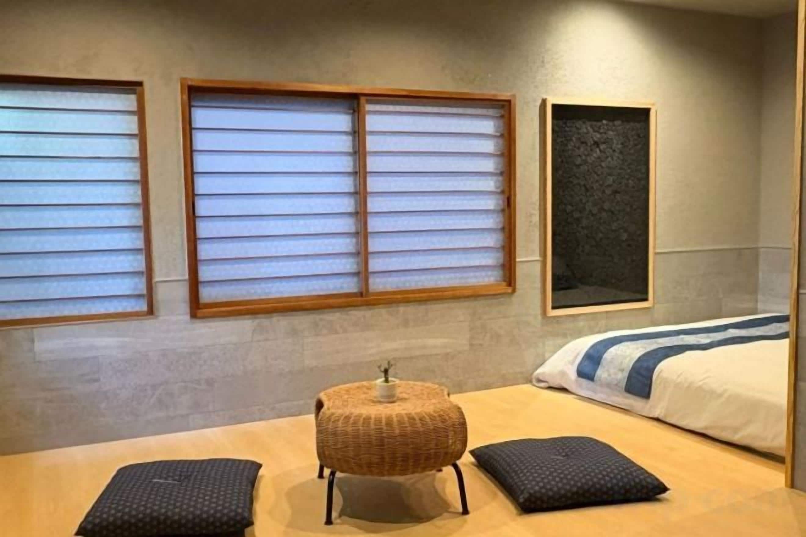 Quarto com cama, duas janelas com persianas, uma mesa pequena com cesta e um travesseiro.