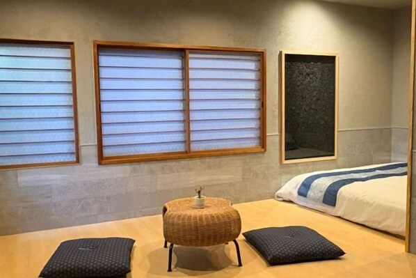 Una habitación con una cama, dos ventanas con persianas, una mesita con una cesta y una almohada.