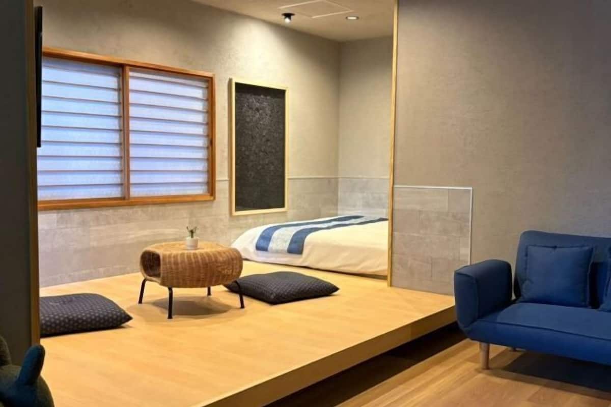 A modern hotel room with a bed, a blue sofa, a small table with a lamp, and a window with blinds.