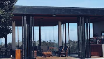 Un área de estar al aire libre techada con sillas y una mesa, ventanas grandes y vista al paisaje.