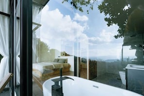 A modern bedroom with a glass wall, a bed, and a bathroom with a bathtub.