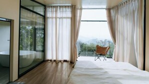 A modern bedroom with a large bed, a glass-enclosed bathroom, and a view of the outdoors.