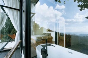 A modern bedroom with a large window, a white bathtub, and a view of the outdoors.