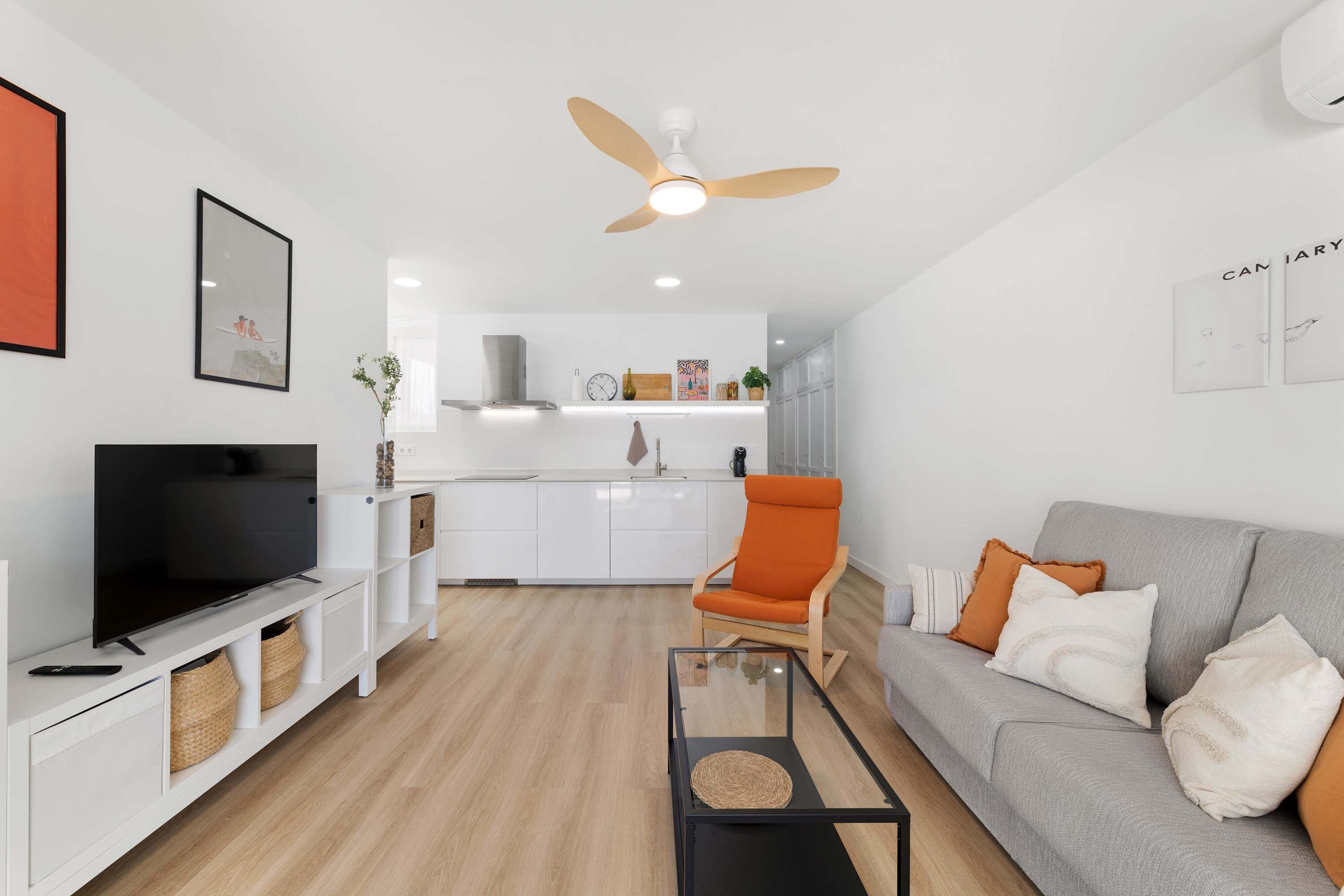 A modern living room with a flat-screen TV, a grey sofa, an orange armchair, and a ceiling fan.