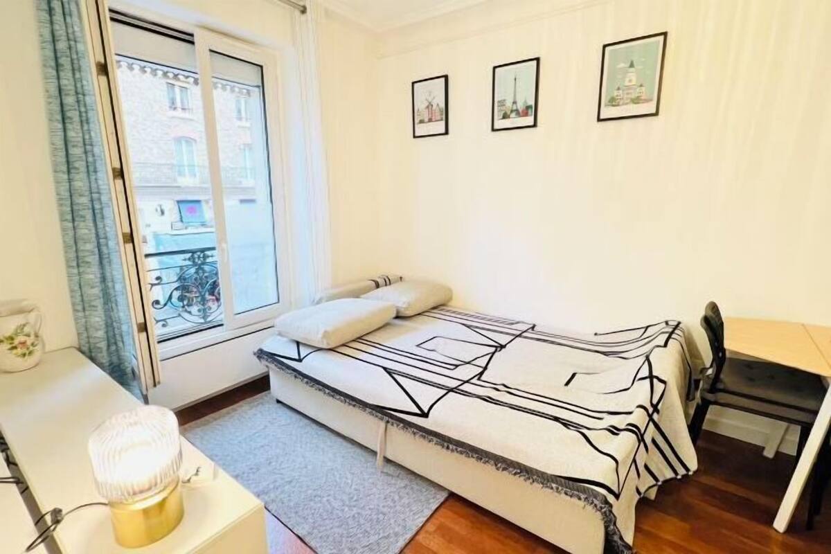 A small bedroom with a bed, a desk, and a chair. There is a window with a view of buildings outside.