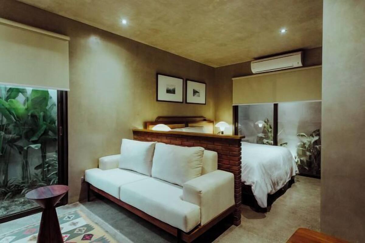 A modern hotel room with a sofa, a bed, a small table, and a large window with plants.
