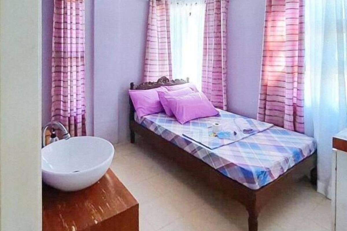 Una cama individual con colcha a cuadros, dos almohadas, cabecero de madera y un lavamanos blanco en un baño con cortinas rosas.