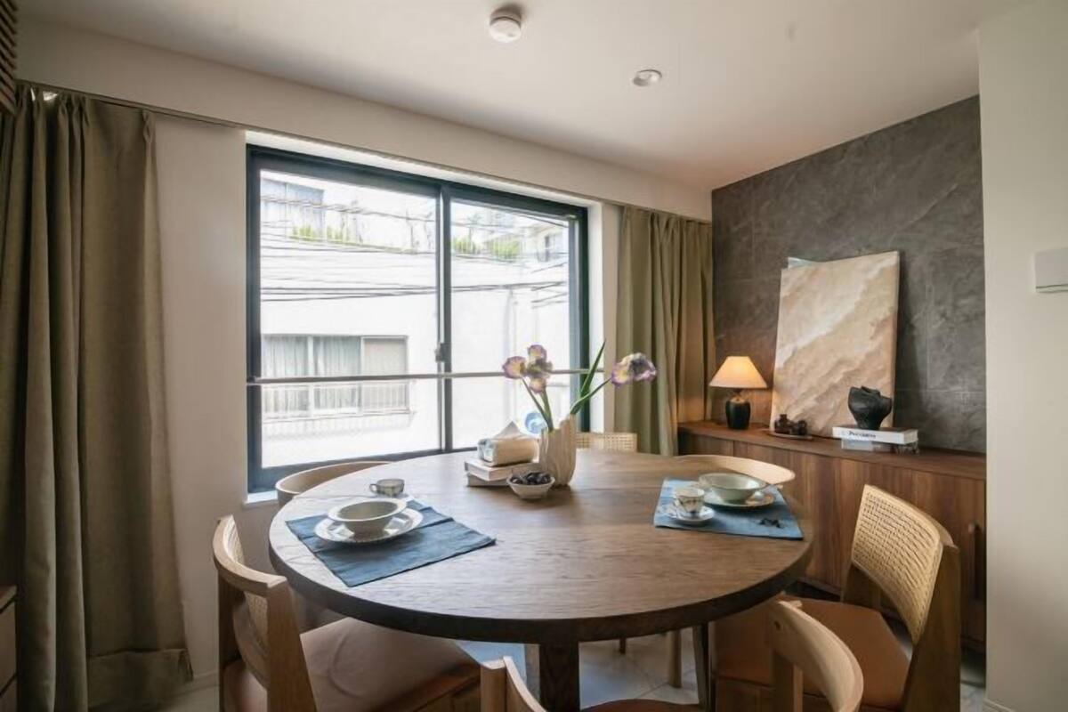 A hotel room with a round dining table, chairs, a vase with flowers, a lamp, and a view of buildings through a large window.